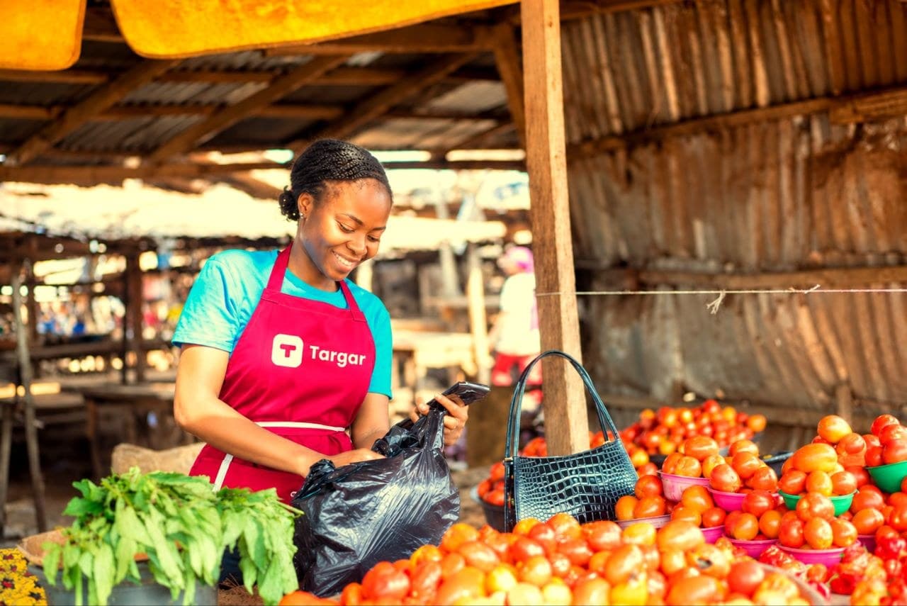 A TARGAR trader packs fresh tomatoes into a bag while completing a payment on her phone